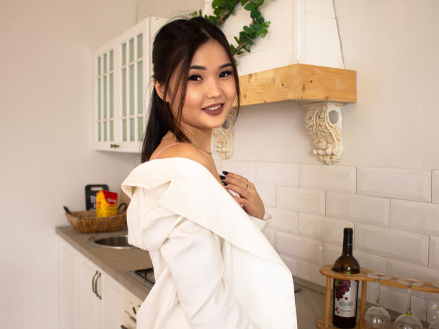TeyaFaber with dark hair, smiling, wearing a white off-shoulder shirt, exposing a small, cute ass, in a kitchen with a wooden shelf and a bottle of wine.