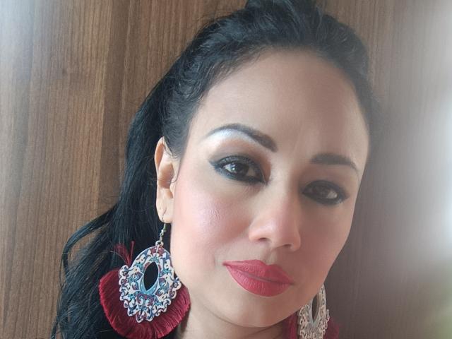SamantaDark with dark eyes, red lipstick, and a smile, wearing a red top and a large, ornate blue and silver earring. Black hair in a ponytail, wooden background.