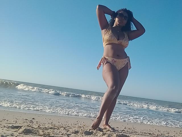 CaroldelaTorre with curly dark hair, wearing a yellow floral bikini, standing on a sandy beach, hands behind head, clear blue sky, small waves in the background.