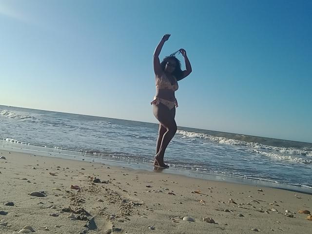 CaroldelaTorre with arms raised, wearing a beige bikini, standing on a sandy beach with gentle waves, clear blue sky, and scattered small rocks in the foreground.