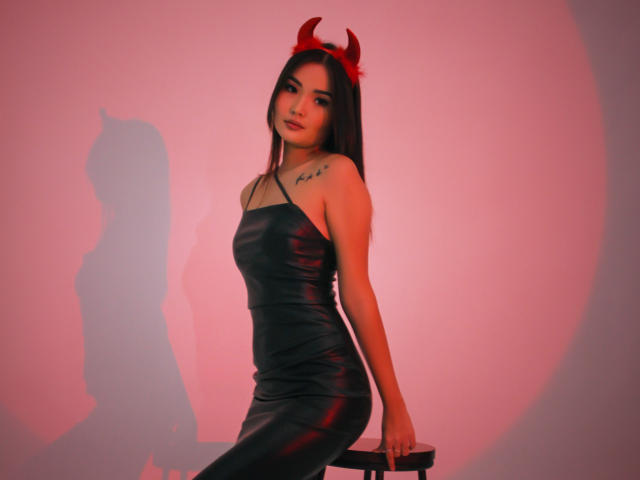 TeyaFaber, con cabello oscuro, cuernos rojos de diablo, vestido de cuero negro, tatuaje en el hombro derecho, sentada en un taburete, iluminación rosa, sombra en la pared, sexy, delgada, linda, segura de sí misma..