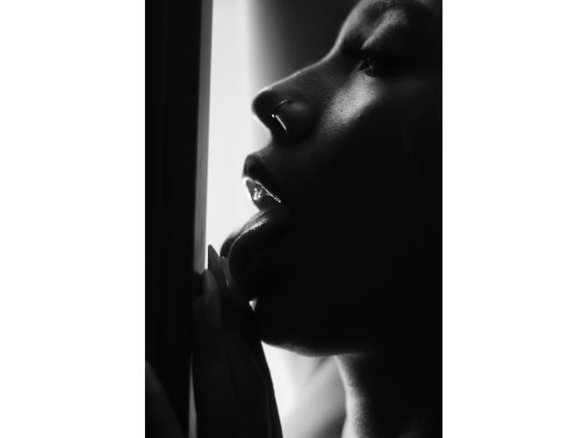 LilithWalker's dark, glossy lips are parted, tongue slightly out, touching a window's edge in a high-contrast black-and-white shot, highlighting her smooth, reflective skin.