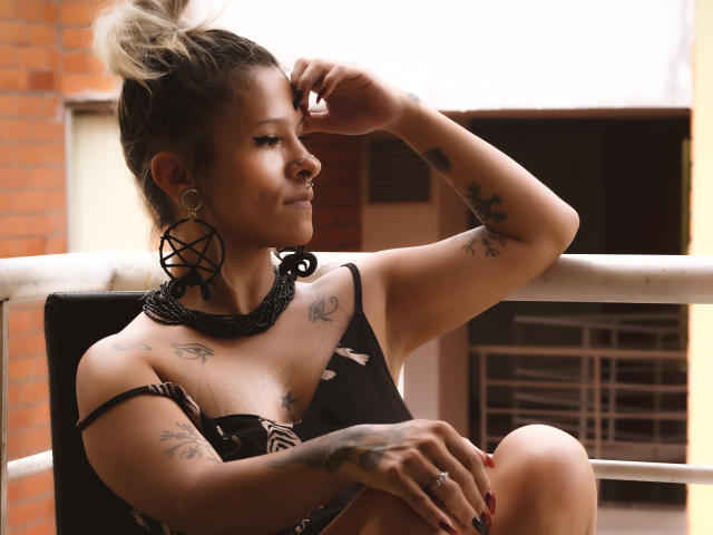 AmelieBlake with tattoos, wearing a black, strappy top, has her blonde hair in a messy bun, large circular earrings, and a black necklace, sitting on a balcony, smirking.