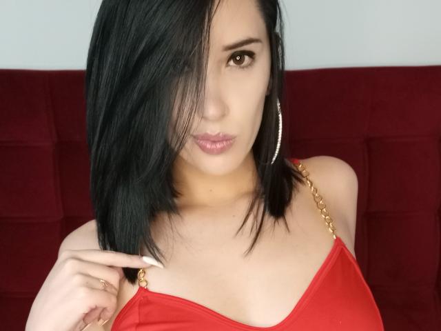 Devorah69 with black hair, red dress, gold hoop earrings, and a finger near her collarbone, looking at the camera with pouty lips. Boobs visible, medium-sized.