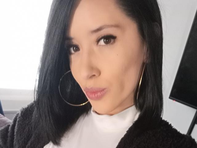 Devorah69 hat mittellanges, schwarzes Haar, braune Augen und rosafarbene Lippen. Sie trägt goldene Ohrringe im Stecker-Stil und einen schwarzen, flauschigen Pullover über einem weißen Rollkragenpullover..