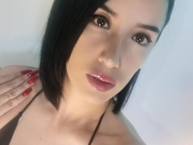 Devorah69 a des lèvres roses brillantes, une coupe de cheveux noire et lisse, des ongles rouges, des yeux marrons et un joli visage. Elle porte un haut marron et a une main sur son épaule..