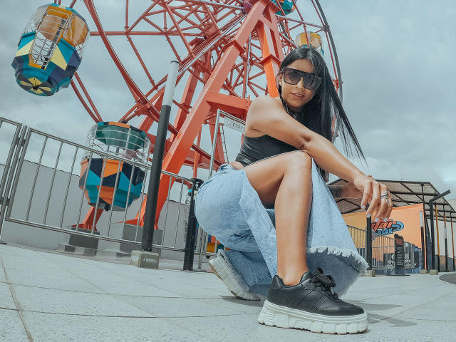 Amberv crouches in front of a red amusement ride, wearing a black tank top, blue jeans, black sneakers, and sunglasses. Her ass is exposed, long black hair down.