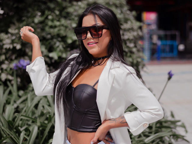 Amberv with black leather boobs, white jacket, black choker, long black hair, black sunglasses, flexing arm, tattoo on hand, purple lipstick, standing outdoors, green foliage in background.