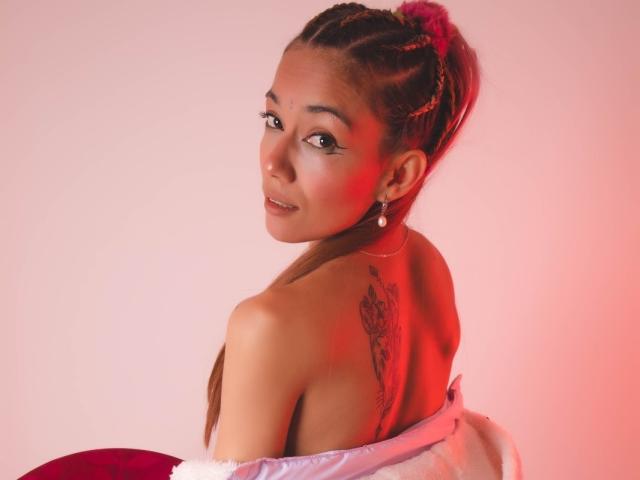 Veroo69 with braided pink hair, looking back, showing a tattoo on her shoulder, wearing a red and white off-shoulder top, smiling with red and pink lighting.