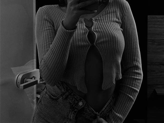 Black and white photo of NutaAlisson. Ribbed, long-sleeve, button-up top with cutout, exposing midriff. Unbuttoned jeans. Hand holding phone, covering face. Mirror reflection. Dark background.