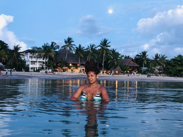 Florinie met donkere huid, krullend haar en kleine borsten, in een blauw-groene bikini, staand in ondiep water, kijkend naar de camera, met palmbomen en een strandhuis op de achtergrond..