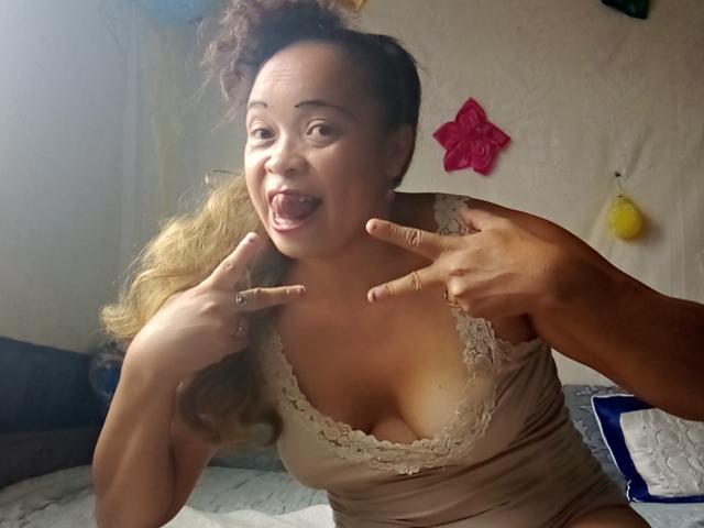 Rahel69 with curly brown hair, tan lace-top tank, medium tits, and tan skin, making double peace signs, mouth open, looking surprised. Hands point near her face. Bedroom setting.