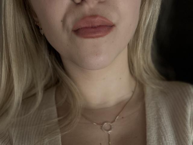 Das Gesicht von SugarrX mit rosafarbenem Lippenstift, leichten Sommersprossen, blonden Haaren, einer kleinen Nase, heller Haut, trägt ein geripptes weißes Oberteil und eine zarte Halskette mit einem runden Anhänger..
