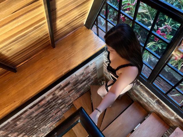TinaYEdward with dark brown hair, black lace bra, and white dress, sitting on wooden stairs, looking down, holding black railing, brick wall, and large window in background.