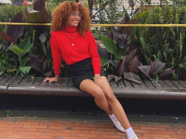 CinthiaWalker, avec ses cheveux bruns bouclés, son pull rouge à col roulé, sa mini-jupe noire, ses chaussettes blanches et ses baskets blanches, assise sur un banc, souriante, montrant ses fesses et ses seins..