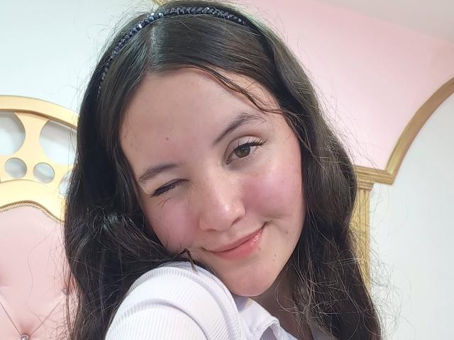 ChloeDulce cligne de l'œil avec un sourire mignon, ses yeux marrons à moitié fermés, portant un bandeau noir dans ses longs cheveux noirs et ondulés. Elle porte une chemise blanche et a l'air séduisante..