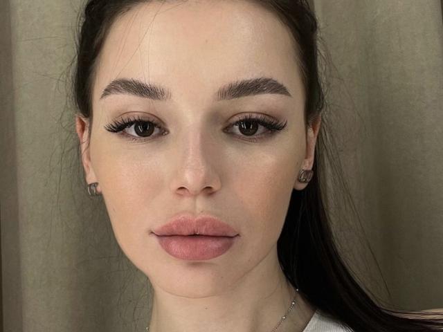 BeckiXHoney's face, showing full lips, dark eyebrows, long eyelashes, and brown eyes, with dark hair in a ponytail, against a beige background. She's wearing small, round earrings.