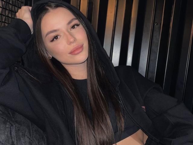 BeckiX, avec de longs cheveux bruns, un sweat à capuche noir et un crop top noir, assise, regardant la caméra, souriant légèrement, avec un anneau de nez noir, un eye-liner noir, des yeux foncés et des lèvres roses..