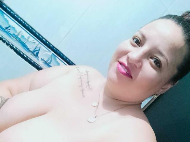 SkayLaraWalker con lápiz labial rosa, cabello oscuro recogido en un moño, sonriendo, sin top, mostrando grandes senos, tatuaje en el hombro derecho, collar y una pared negra detrás de ella..