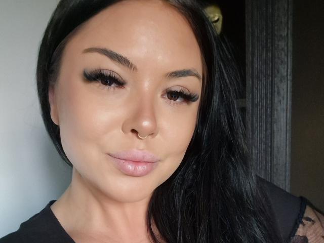 DarkElf tiene el cabello negro brillante, ojos marrones, pestañas gruesas, un piercing en la nariz y labios rosados y brillantes. Lleva una blusa negra transparente y está mirando a la cámara..