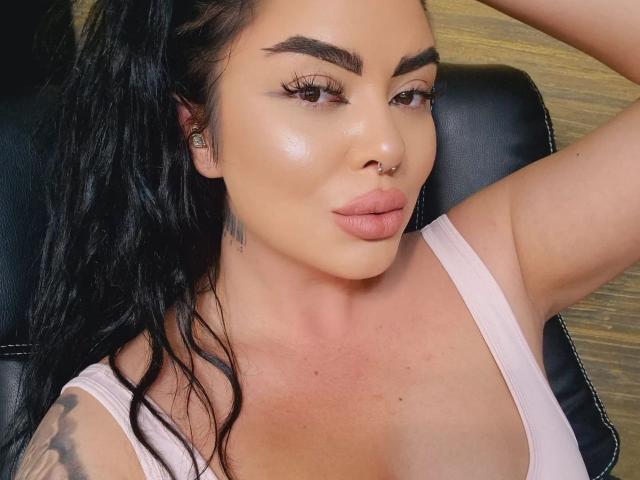 Elfo oscuro con piel brillante, cejas gruesas, cabello negro largo, aro en la nariz y camiseta rosa, mostrando labios carnosos y un brazo tatuado, sentado sobre cuero negro..