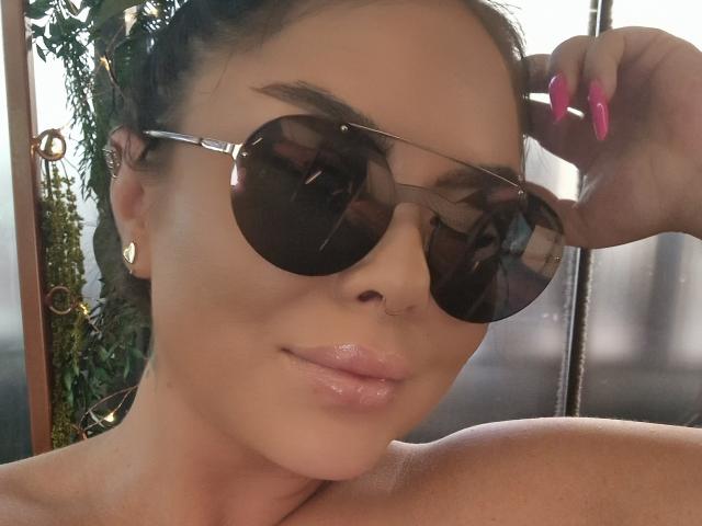 DarkElf with glossy pink lips, shiny skin, and pink nails, wearing round sunglasses, touching her nose with a finger, and a nose ring. Her brown hair is pulled back.