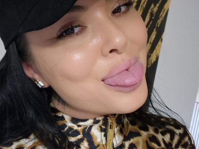 Il viso di DarkElf, con una lingua rosa e lucida che sporge, indossa un cappello nero e una giacca con stampa leopardo e una cerniera dorata. Ha un piercing al naso e orecchini lucidi..