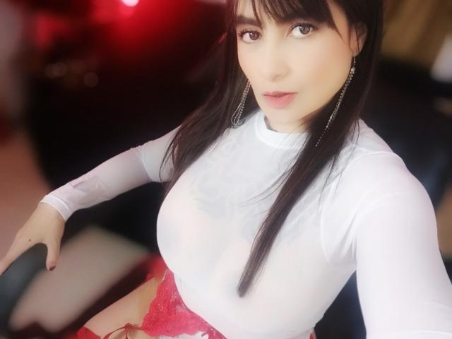 BelleLoren with long black hair, white sheer top showing her tits, pink lace skirt, and dangling earrings, smiling slightly. Red light in the background.