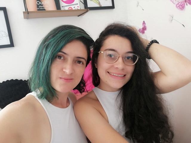 LunaVenus with green hair and LunaVenus with black hair, both in white tank tops, smiling at the camera, glasses on, arm around each other, white wall background.