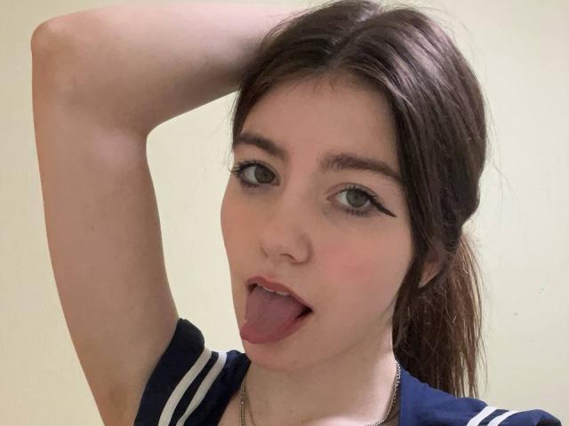 VinnaSkyHott has brown hair, green eyes, and is sticking out her tongue with her arm raised. She's wearing a navy blue shirt with white stripes.