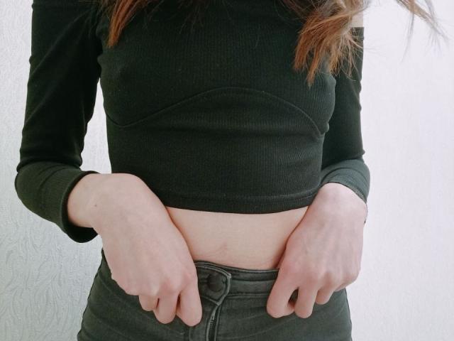 WollyMolly's hands are gripping the waistband of her green pants, exposing her pale midriff, wearing a black ribbed crop top, with brown hair falling.