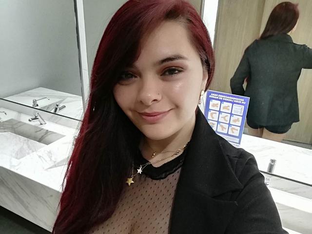 katherinLatin with red hair, smiling, wearing black coat over sheer polka-dot top, showing small tits, gold necklace, mirror reflection showing back, standing in bathroom.