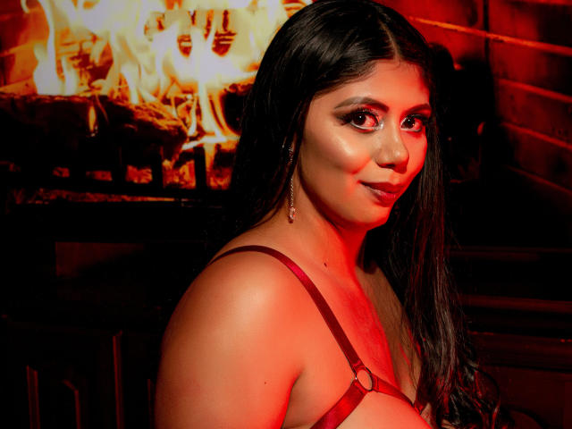 CloeSweety with long dark hair, red eyes, and glossy makeup, wearing a red strap harness, looking over her shoulder with a slight smile, in a red-lit setting.