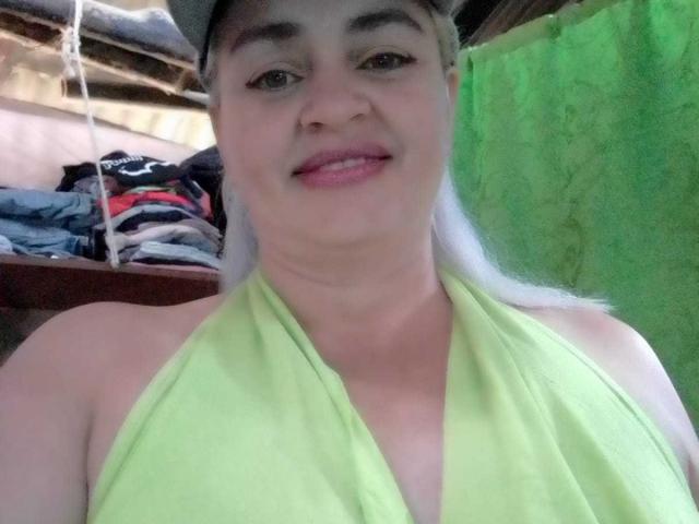 SalomeAriza has light skin, green halter top, and a slight smile. She's wearing a cap, with visible tits under the top, and a green curtain in the background.
