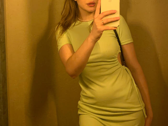 NutaAlisson takes a mirror selfie. She wears a tight, green dress with short sleeves and a cut-out side. Her right hand holds a phone, partially covering her face.