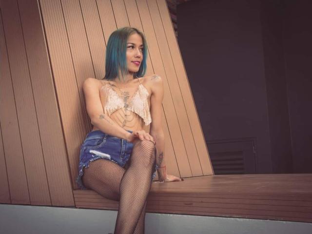 Veroo69 with blue hair, lace-up top, denim shorts, fishnet stockings, sitting on wooden ledge, showing her ass and tattooed arm, smiling, looking to the side.