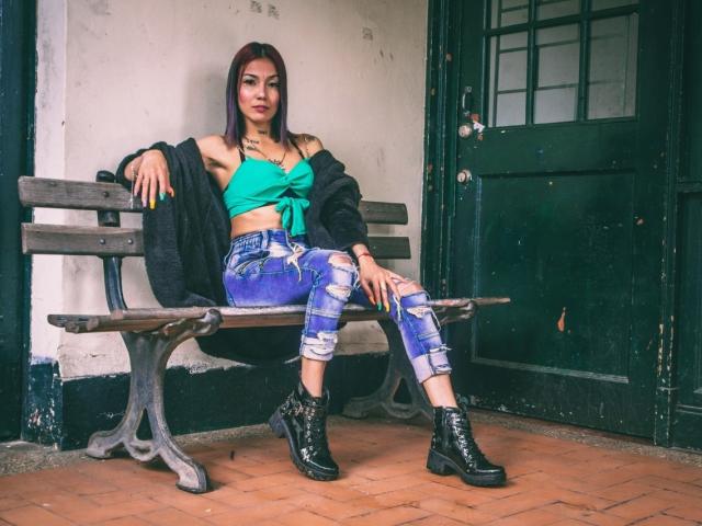 Veroo69 with purple hair, green top, ripped blue jeans, black boots, and black jacket, sitting on a wooden bench, hands on knees, looking at camera.