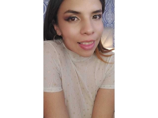 VioletaStone has tan skin, brown eyes with eyeliner, pink lips, and a small smile. She's wearing a sheer, white, dotted top with hoop earrings.