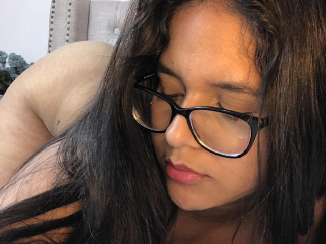 Megan Brownish, avec de longs cheveux noirs, des lunettes noires et du rouge à lèvres rose, les yeux fermés, regarde vers le bas. Ses fesses sont partiellement visibles en arrière-plan. Son visage est au premier plan..