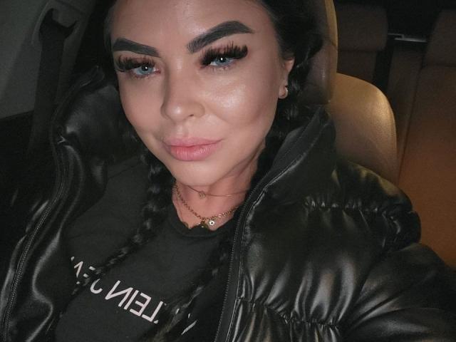 DarkElf with blue eyes, dark eyebrows, long black eyelashes, and braided hair, wearing a black leather jacket, sitting in a car, smiling slightly, with a subtle sheen on her face.