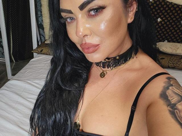 Elfa oscura con piel brillante, cabello negro, cejas gruesas y labios rosados. Lleva un collar negro, un collar dorado y un top de tirantes negros, mostrando un tatuaje en su brazo derecho..