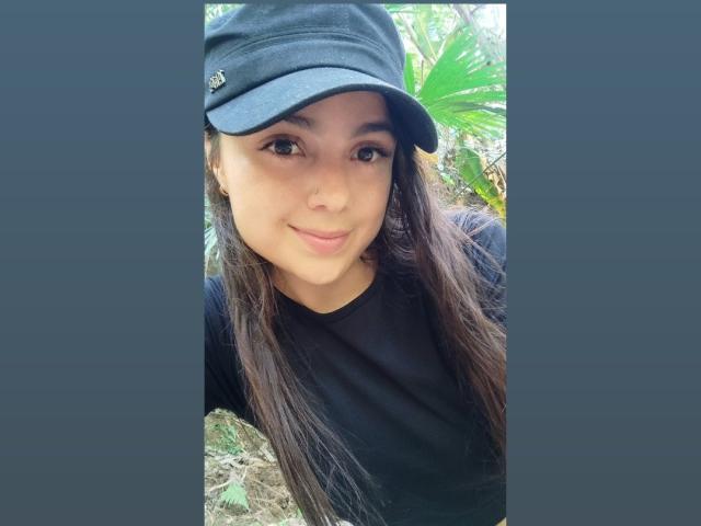 HeidyMillerr has long brown hair, a cute smile, and brown eyes, wearing a black cap and black shirt. She's outdoors with green leaves in the background.