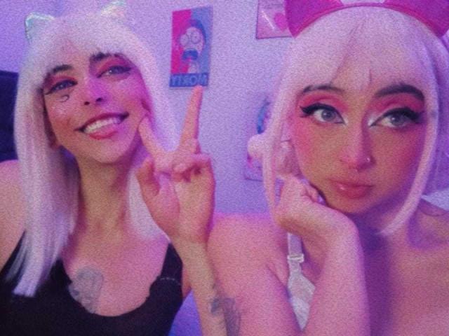 TheLesbianGame, com orelhas de gato brancas, top preto e tatuagens, fazendo um sinal de paz, tem sombra rosa nos olhos, um piercing no nariz e está ao lado de outra TheLesbianGame com um chapéu rosa, com uma expressão emburrada..