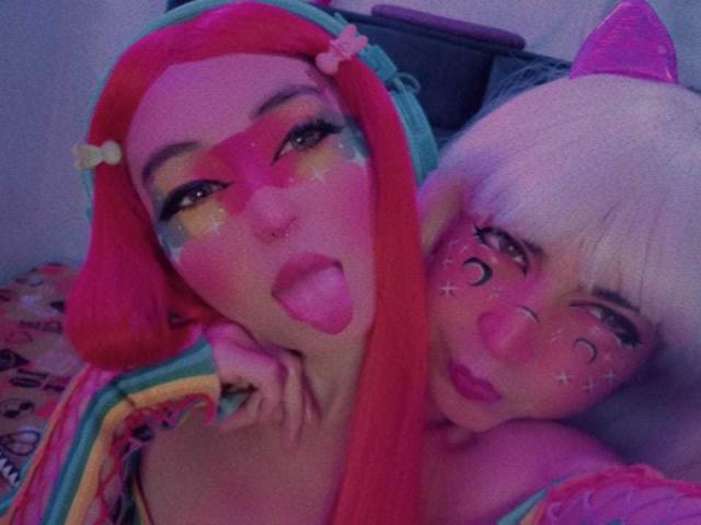 Deux femmes avec un maquillage coloré, TheLesbianGame à gauche avec des cheveux roux, la langue tirée, et TheLesbianGame à droite avec des cheveux blancs, toutes deux avec des visages scintillants et des t-shirts arc-en-ciel..