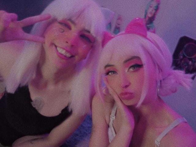 TheLesbianGame avec des cheveux blancs, un haut noir, et TheLesbianGame avec des cheveux roses, un haut blanc et des oreilles de chat roses, toutes deux faisant des signes de "V" et souriant, dans une pièce éclairée en rose..