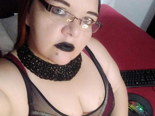 Andrea Sharlin, usando óculos, batom preto e um colar de contas pretas, vestindo uma blusa preta transparente que revela seus seios grandes e o colo, sentada em frente a uma cadeira vermelha..