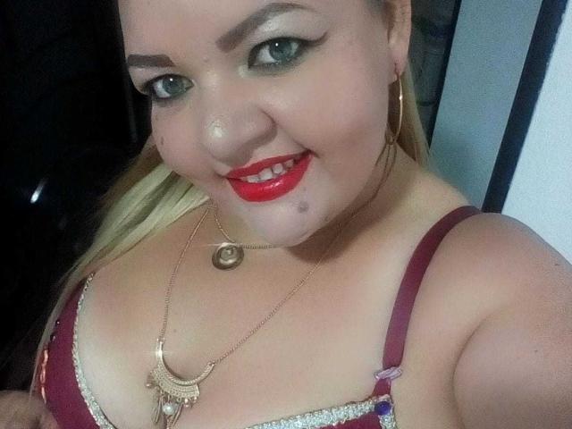 Andrea Sharlin, com cabelo loiro, batom vermelho e sombra verde, vestindo um sutiã marrom com um colar dourado, tem esperma no queixo, sorrindo para a câmera..