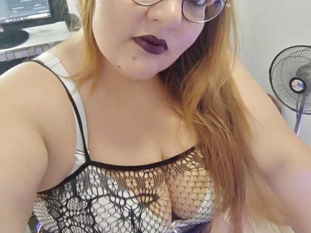 Andrea Sharlin, avec ses longs cheveux roux, ses lunettes et son rouge à lèvres foncé, porte un haut en dentelle noire laissant entrevoir ses grandes seins et un soutien-gorge blanc, et elle est assise dans une pièce avec un ventilateur..