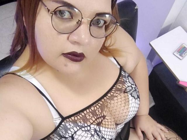AndreaSharlin con gafas, lápiz labial oscuro, top de rejilla blanco y negro que muestra sus pechos, piel clara, cabello castaño rojizo, sentada, mirando hacia arriba a la cámara, en una silla..