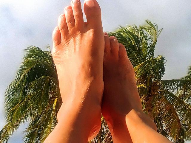 LylyFrench's toes are up in the air, with her soles facing forward, showing slight moisture on her tan skin. Palm leaves are in the background.